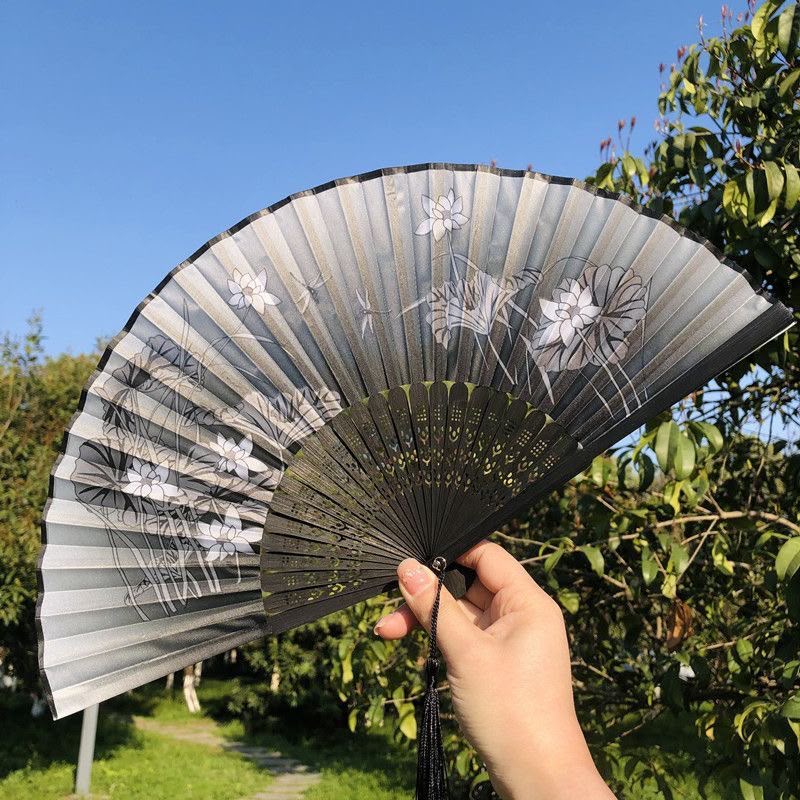 A hand holds a folding hand fan outdoors. The fan has a black frame and translucent gray-white paper panels decorated with white floral and dragonfly illustrations.